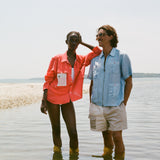 Man and woman standing ankle-deep in the water on a beach. The man wears a light blue zip-up shirt with red and green buttons and four front pockets. The woman wears a bright red long-sleeve shirt over a white swimsuit.