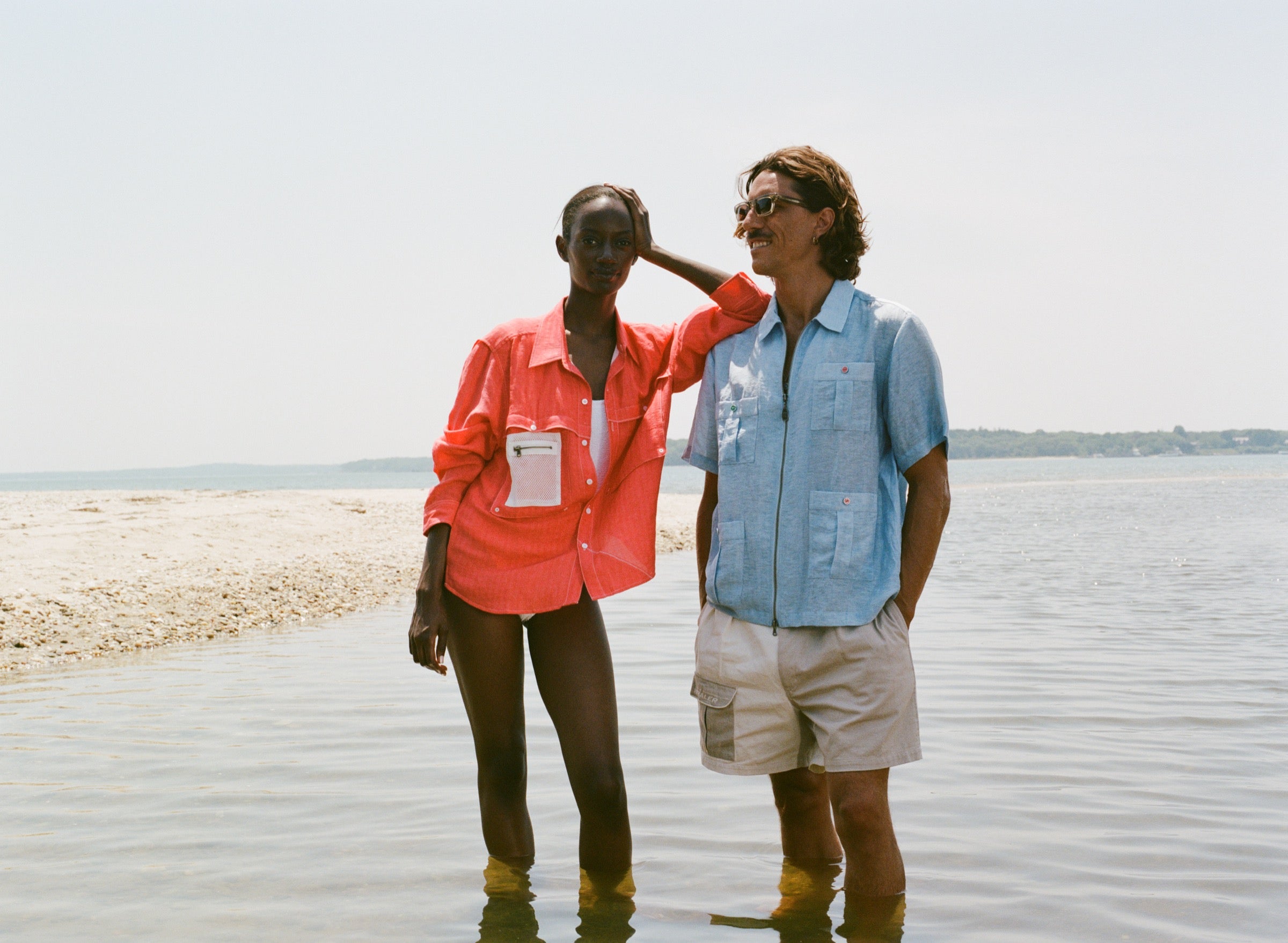 Man and woman standing ankle-deep in the water on a beach. The man wears a light blue zip-up shirt with red and green buttons and four front pockets. The woman wears a bright red long-sleeve shirt over a white swimsuit.
