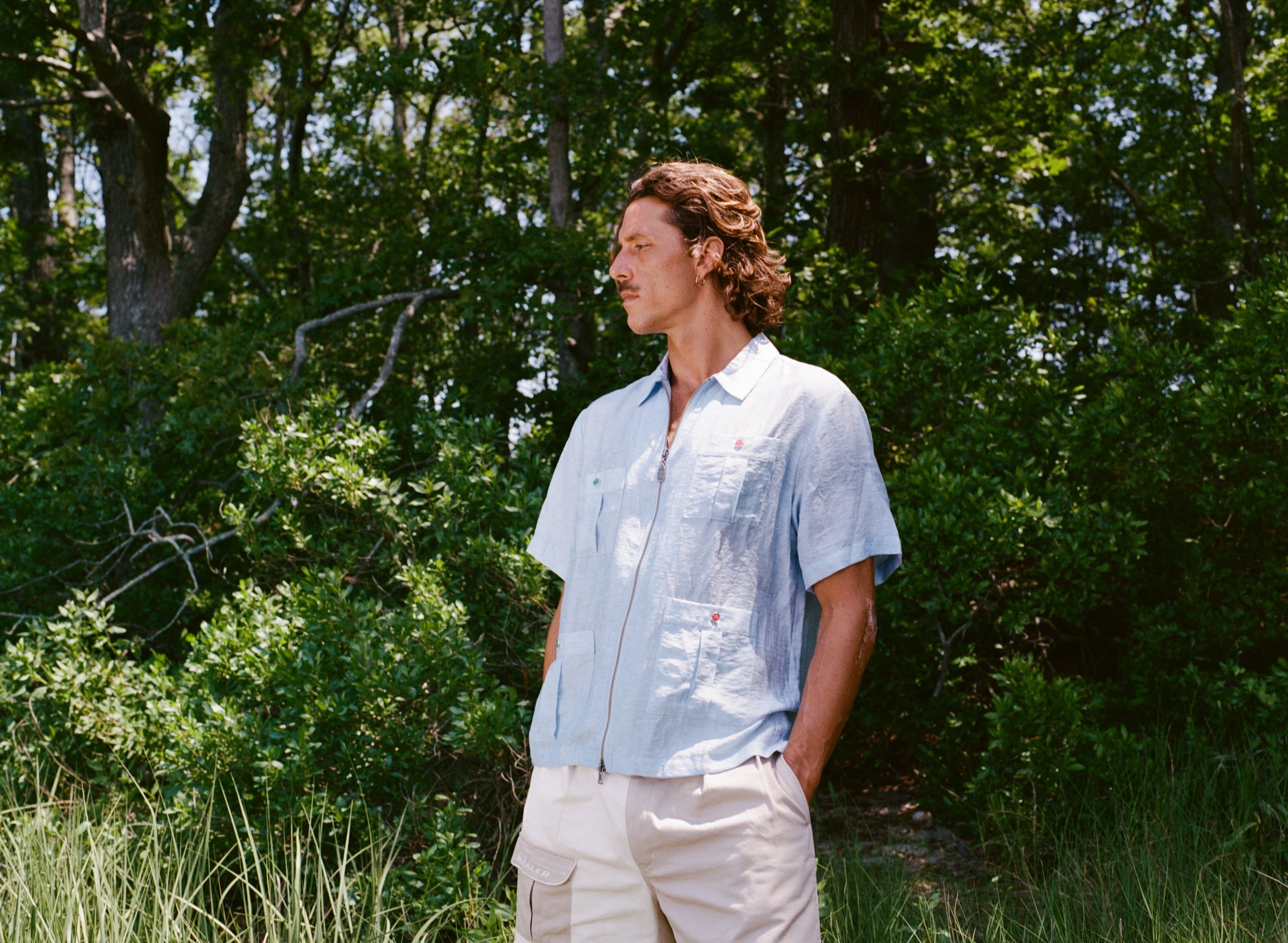 Man stands in grass wearing a light blue zip-up shirt with four front pockets, each fastened with red or green buttons, and khaki shorts with the word 'Whaler' embroidered on the pocket.