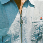 Close-up of a light blue zip-up shirt with four front pockets. Each pocket is fastened with a green or red button. The zipper pull is shaped like a boat and engraved with 'BW'.