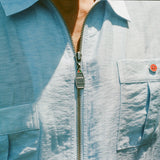 Close-up of a light blue zip-up shirt with four front pockets. Each pocket is fastened with a green or red button. The zipper pull is shaped like a boat and engraved with 'BW'.