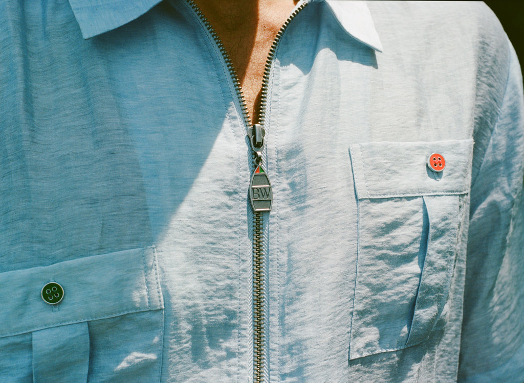 Close-up of a light blue zip-up shirt with four front pockets. Each pocket is fastened with a green or red button. The zipper pull is shaped like a boat and engraved with 'BW'.