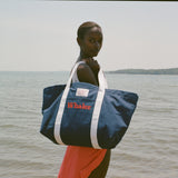 Woman standing in shallow water, holding the navy 'Whaler' canvas tote bag with white handles over her shoulder.