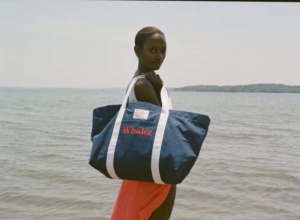 Woman standing in shallow water, holding the navy 'Whaler' canvas tote bag with white handles over her shoulder.