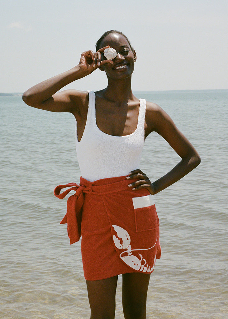 woman at beach wearing white one piece bathing suit and red skirt, smiling and holding a shell up to her eye 