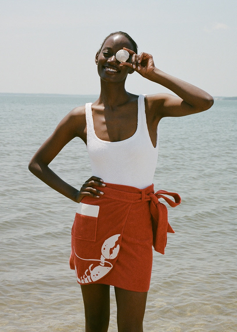 woman at beach wearing white one piece bathing suit and red skirt, smiling and holding a shell up to her eye 
