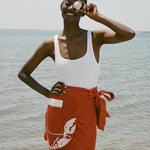 woman at beach wearing white one piece bathing suit and red skirt, smiling and holding a shell up to her eye 
