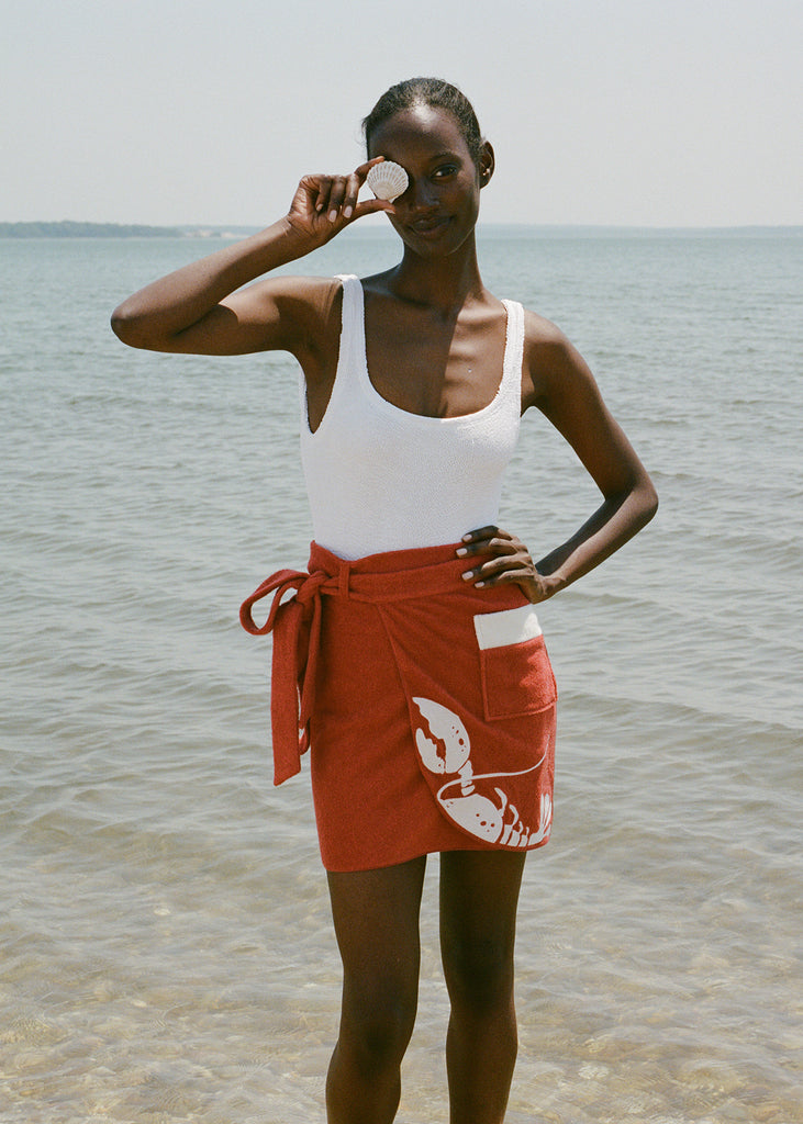 woman at beach wearing white one piece bathing suit and red skirt, smiling and holding a shell up to her eye 