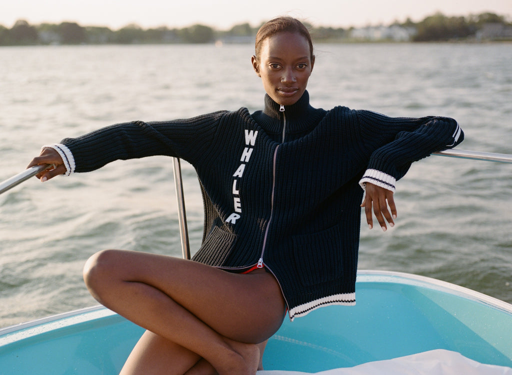 Woman sitting at the back of a boat, wearing a navy zip-up cardigan with vertical white 'WHALER' text and white-striped cuffs.