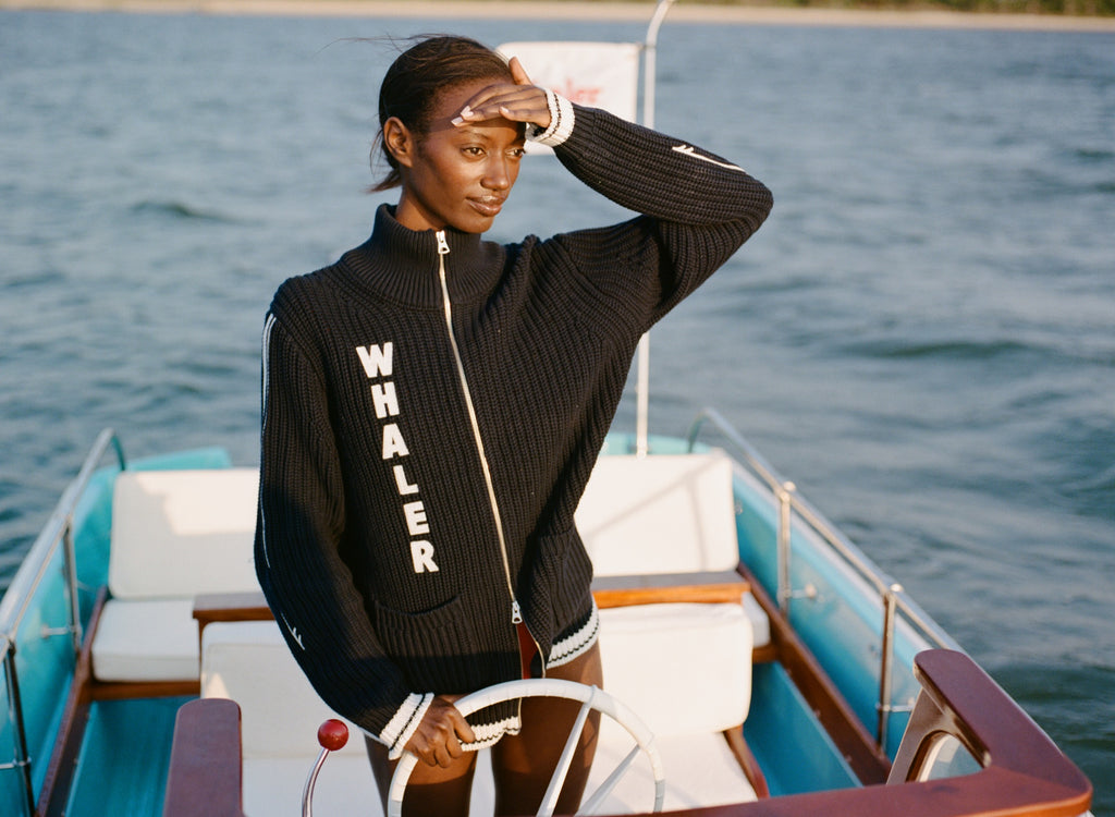 Woman standing at the helm of a boat, wearing a navy zip-up cardigan with vertical white 'WHALER' lettering and white stripes down the sleeves.
