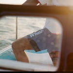 Man standing on a boat wearing navy board shorts with white 'Whaler' text on the leg and contrast stitching. Seen through a window.