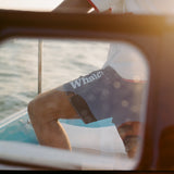 Man standing on a boat wearing navy board shorts with white 'Whaler' text on the leg and contrast stitching. Seen through a window.
