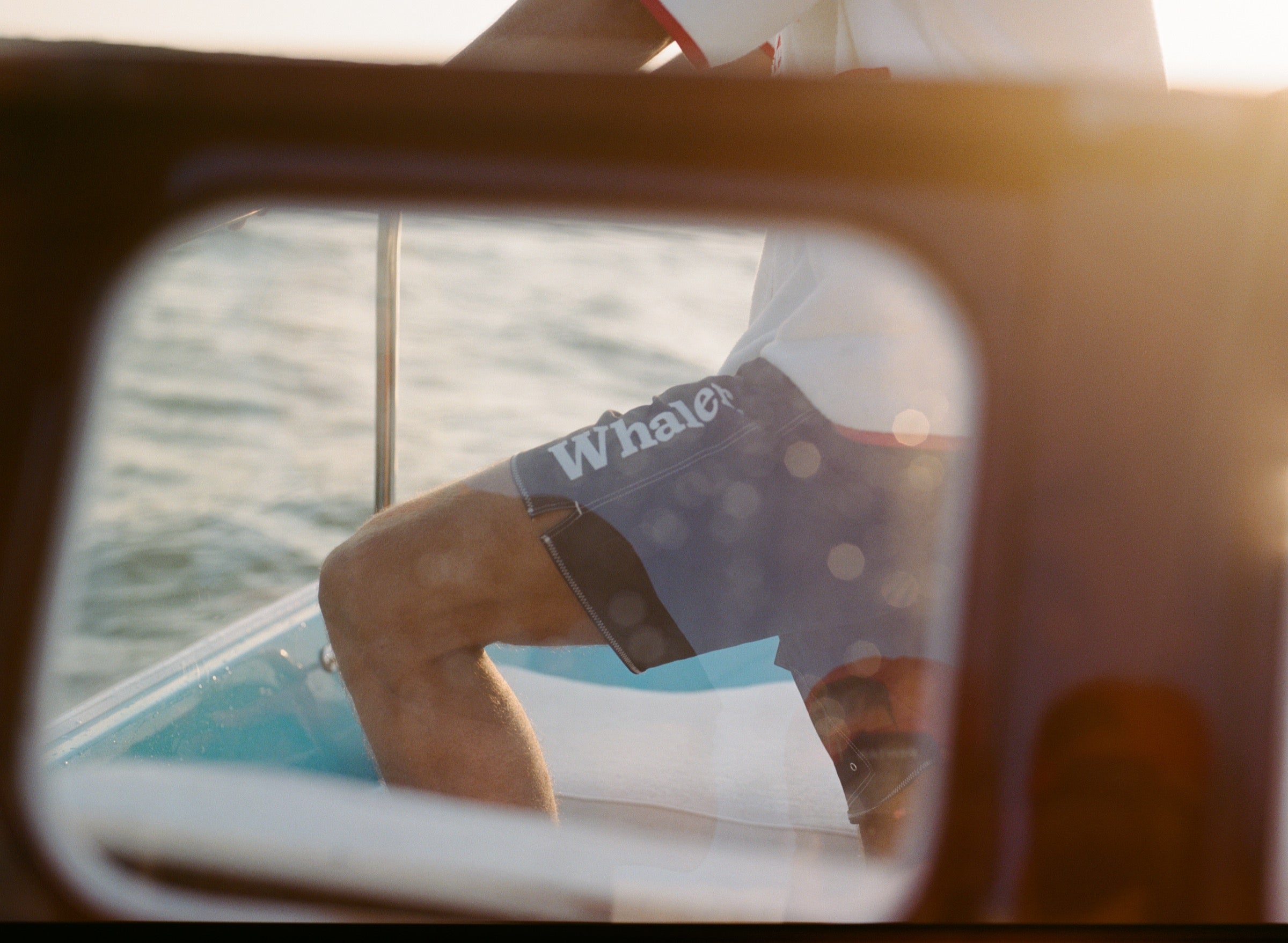 Man standing on a boat wearing navy board shorts with white 'Whaler' text on the leg and contrast stitching. Seen through a window.