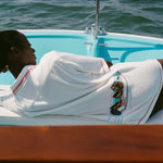 Woman lying on a boat, wearing the white terry 'Boston Whaler' Cabana with a boat embroidery on the back. Her back is to the camera.