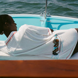 Woman lying on a boat, wearing the white terry 'Boston Whaler' Cabana with a boat embroidery on the back. Her back is to the camera.