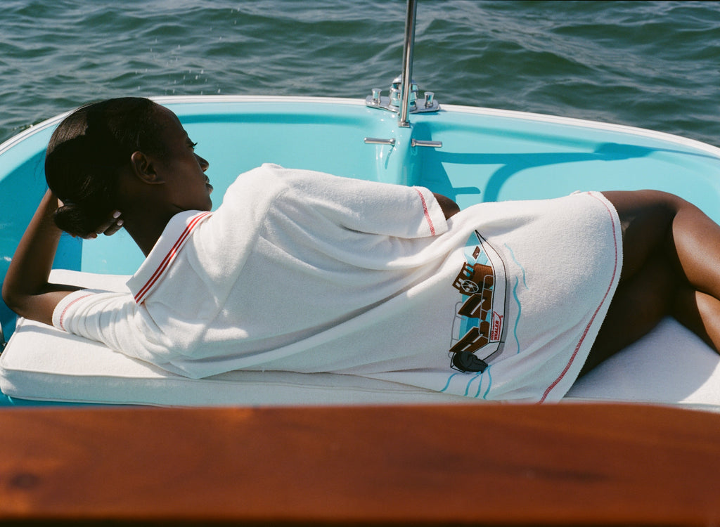 Woman lying on a boat, wearing the white terry 'Boston Whaler' Cabana with a boat embroidery on the back. Her back is to the camera.