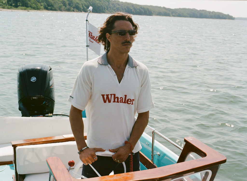 Man with sunglasses on standing at the helm of a boat, wearing a white 'Whaler' polo shirt with red text and a black-and-white striped collar. 