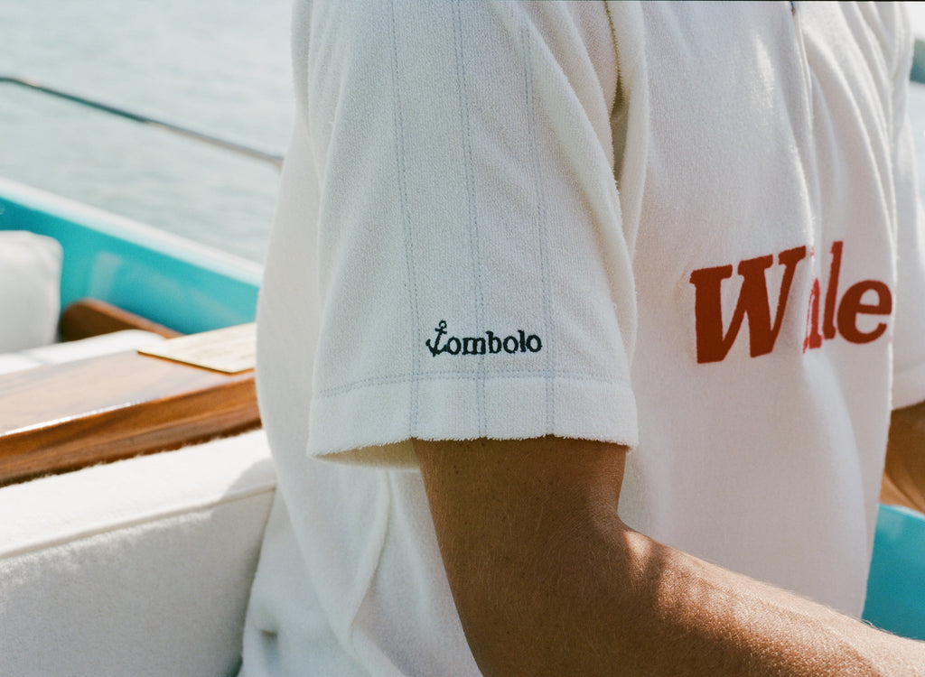 Close-up of a white terry polo shirt showing the left sleeve with black 'Tombolo' logo embroidery and part of the red 'Whaler' text across the chest.