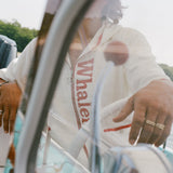 Man wearing white 'Whaler' beach towel top, seen steering a boat. 