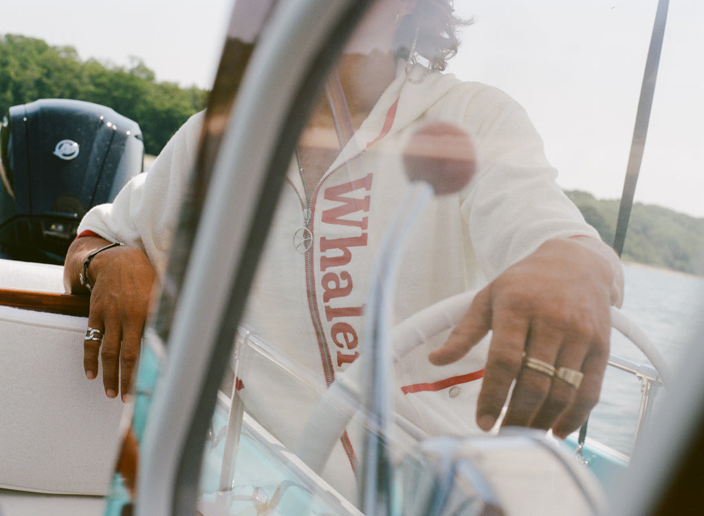 Man wearing white 'Whaler' beach towel top, seen steering a boat. 