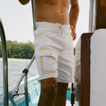 Close-up of a man wearing white board shorts with beige-trimmed cargo pockets and a drawstring waist, standing on a boat.
