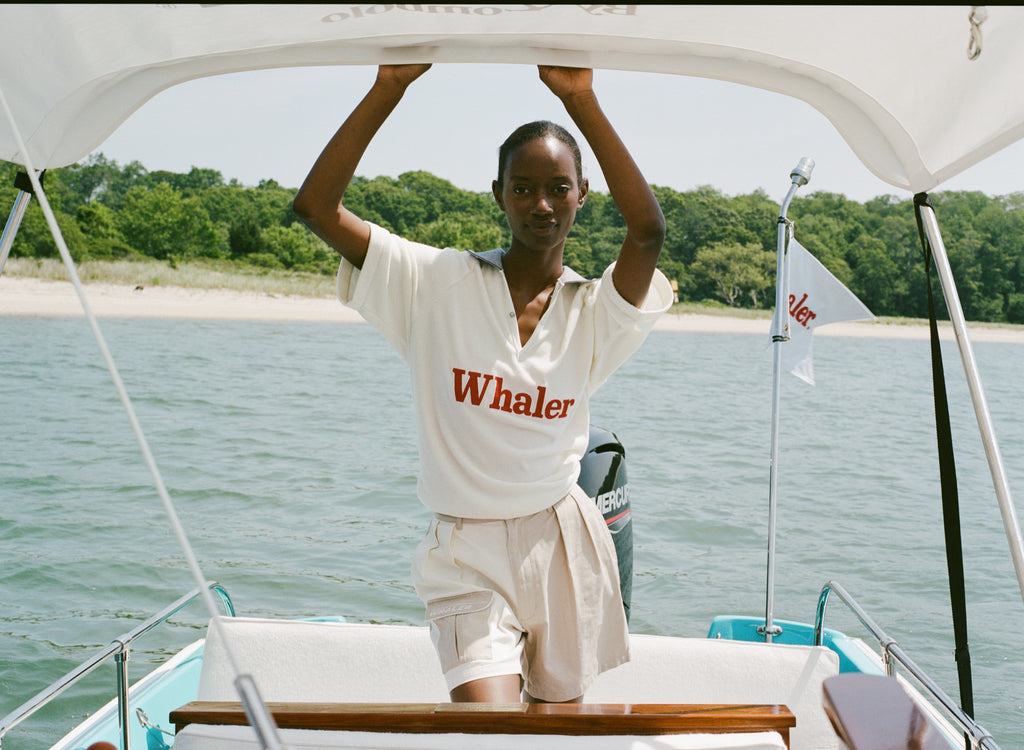 Woman standing on a boat, holding onto the canopy frame. She wears a white 'Whaler' polo shirt with red text and khaki and off-white pleated shorts. A white 'Whaler' flag waves behind her.