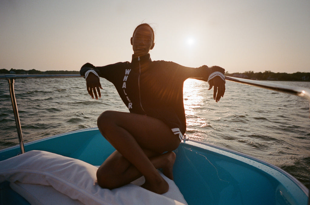 Woman sitting at the back of a boat, wearing a navy zip-up cardigan with vertical white 'WHALER' text and white-striped cuffs.