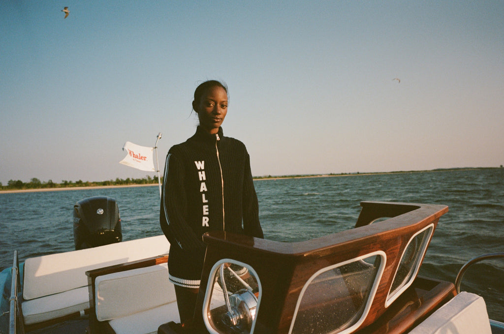 Woman standing at the helm of a boat, wearing a navy zip-up cardigan with vertical white 'WHALER' lettering and white stripes down the sleeves.