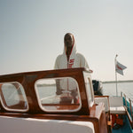 Woman wearing white hooded beach towel top with red trim and vertical 'Whaler' text, standing at the wheel of a boat. 