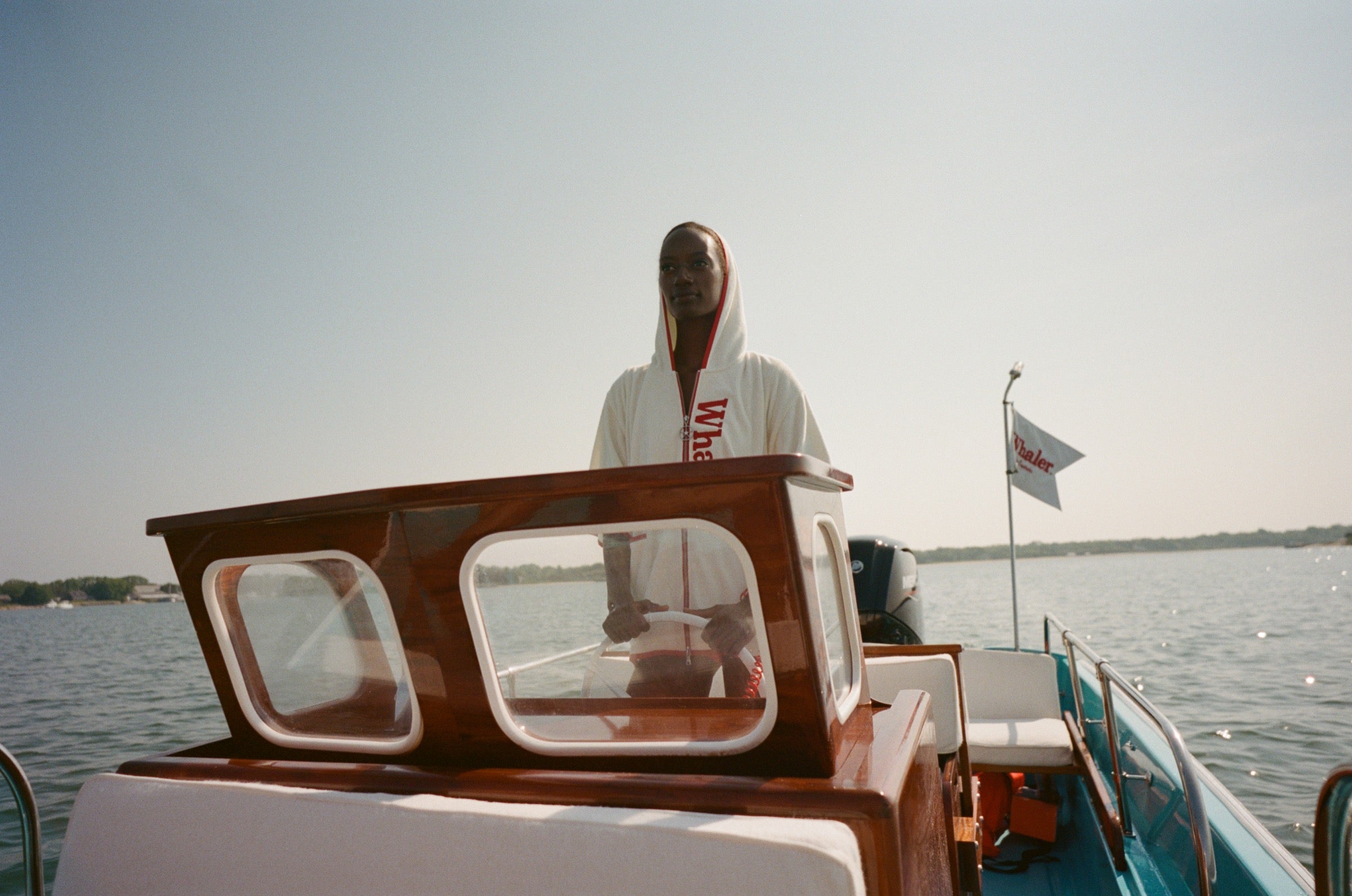 Woman wearing white hooded beach towel top with red trim and vertical 'Whaler' text, standing at the wheel of a boat. 