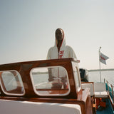 Woman wearing white hooded beach towel top with red trim and vertical 'Whaler' text, standing at the wheel of a boat. 