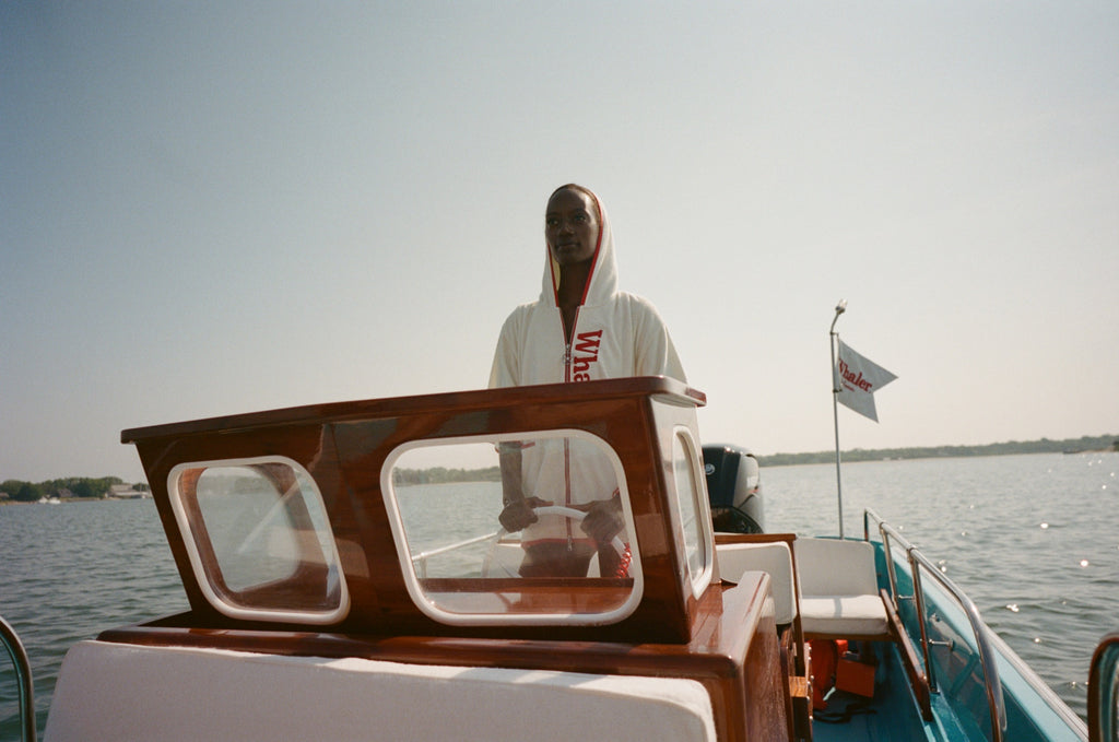 Woman wearing white hooded beach towel top with red trim and vertical 'Whaler' text, standing at the wheel of a boat. 