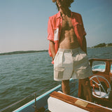 Man wearing two-tone pleated shorts in beige and off-white, featuring a flap pocket on the left with white 'Boston Whaler' embroidery. He wears a red 'Boston Whaler' fishing shirt unbuttoned on top.