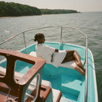 Woman lying on a boat, wearing the white terry 'Boston Whaler' Cabana with a boat embroidery on the back. Her back is to the camera.