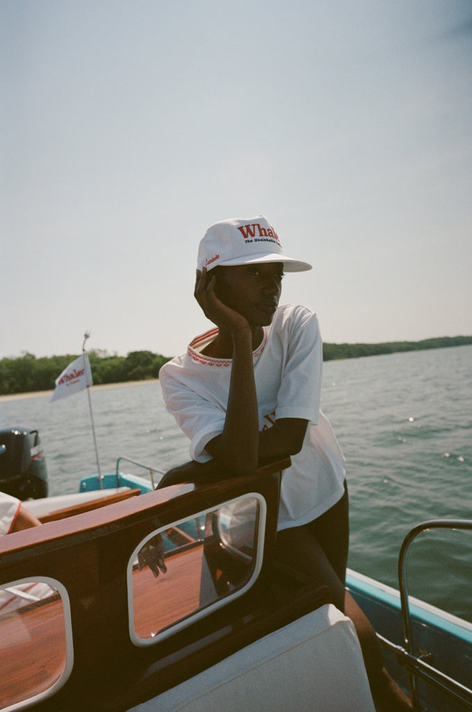 Woman leaning on the cabin of a boat wearing a white five-panel cap with red embroidered text reading 'Whaler' and smaller text reading 'The Unsinkable Legend'. A small red 'Tombolo' logo is visible on the side of the hat. She is also wearing a matching white 'Boston Whaler' shirt with red stitching at the collar.