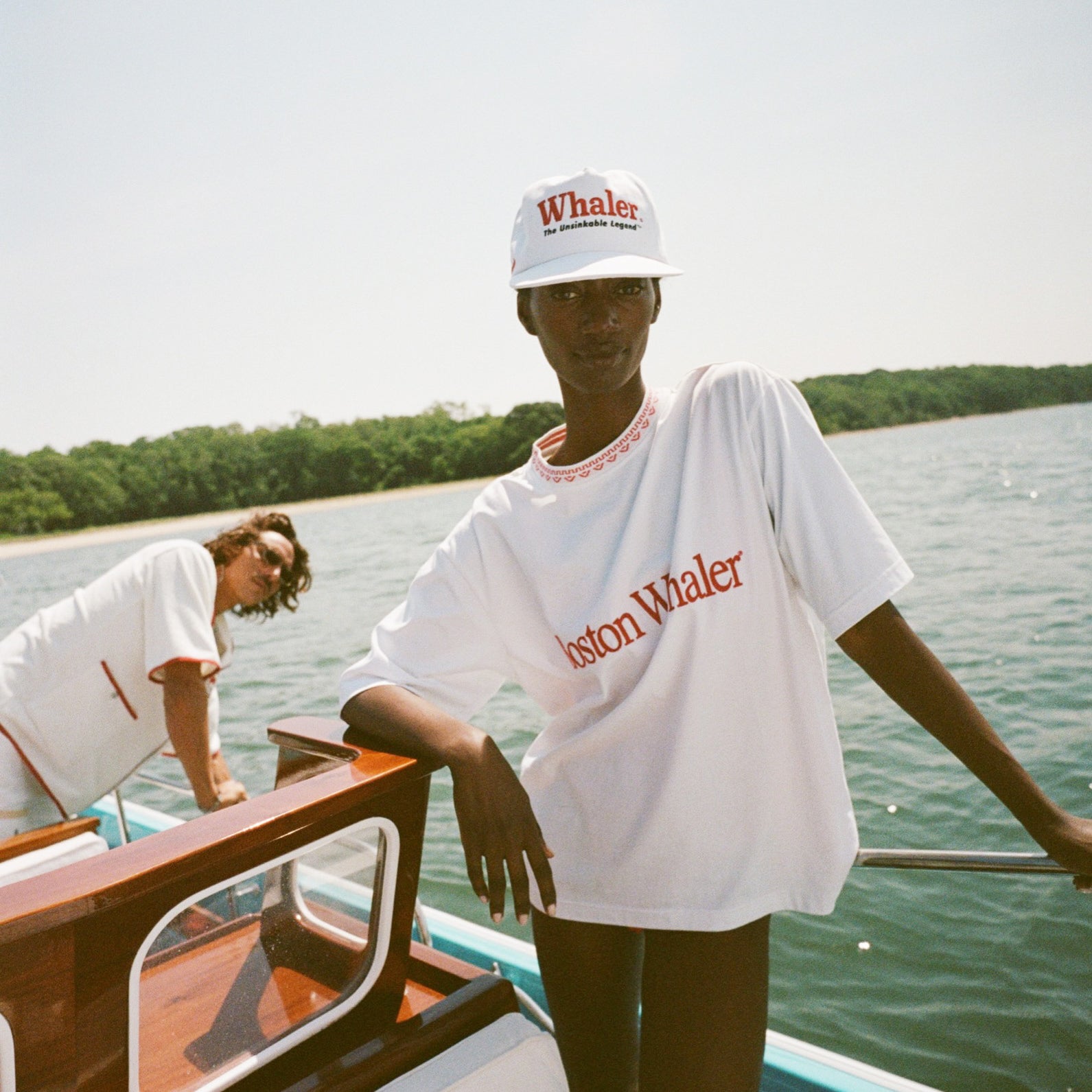 Woman on a boat wears a white 'Boston Whaler' T-shirt with red trim and a white 'Whaler' cap. Behind her, a man wears a white 'Whaler' cabana shirt with red accents.