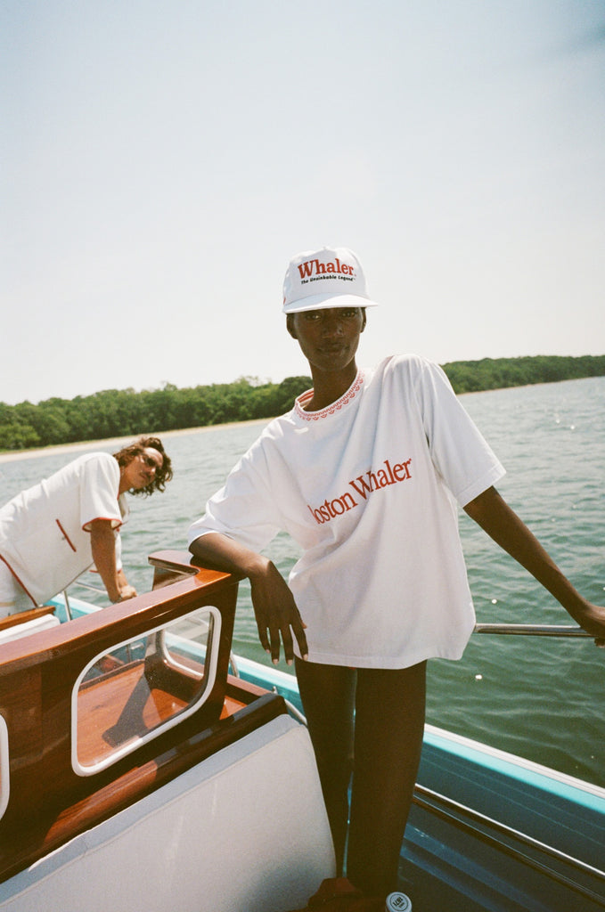 Woman on a boat wears a white 'Boston Whaler' T-shirt with red trim and a white 'Whaler' cap. Behind her, a man wears a white 'Whaler' cabana shirt with red accents.