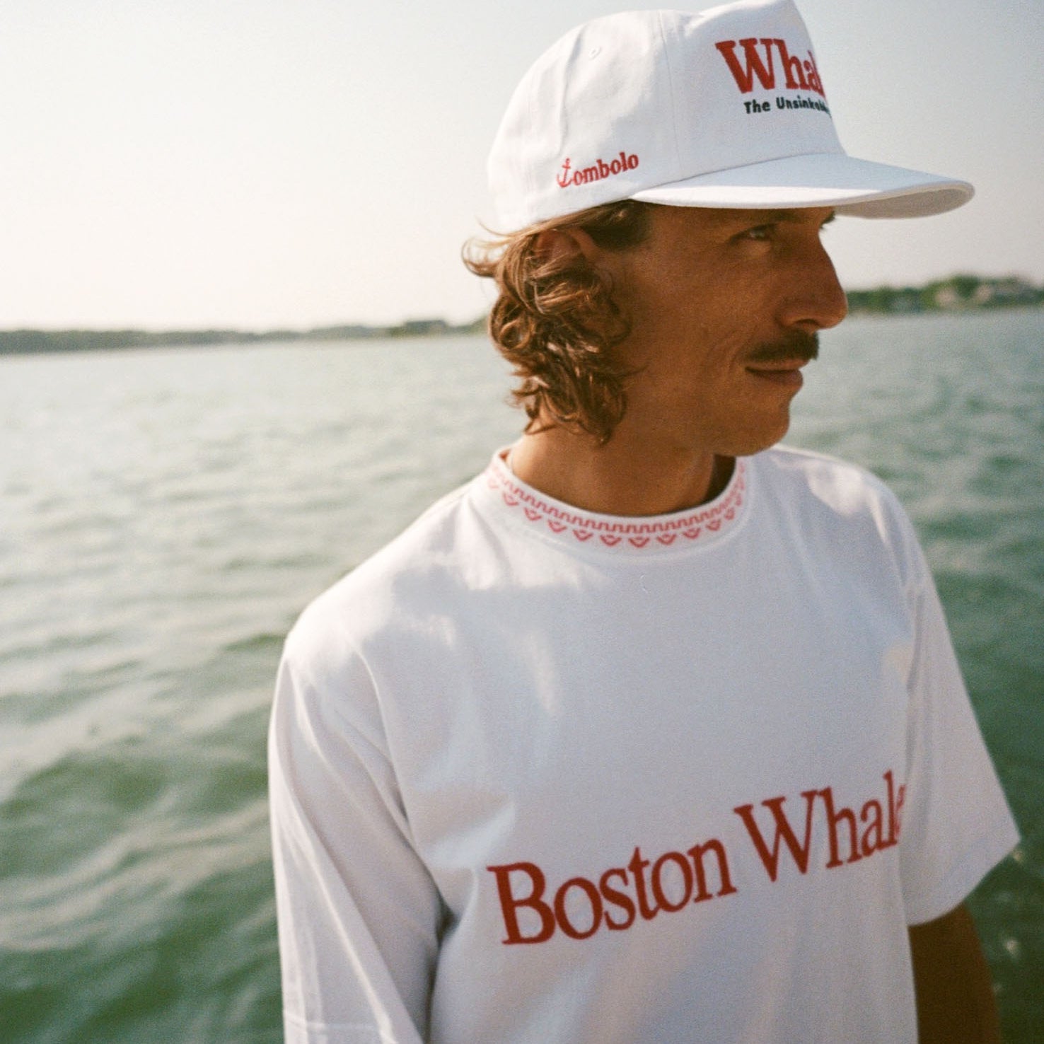 Man on a boat wearing a white 'Whaler' cap with red and black embroidery and a white T-shirt with 'Boston Whaler' in red text. The side of the cap reads 'Tombolo' in red. 