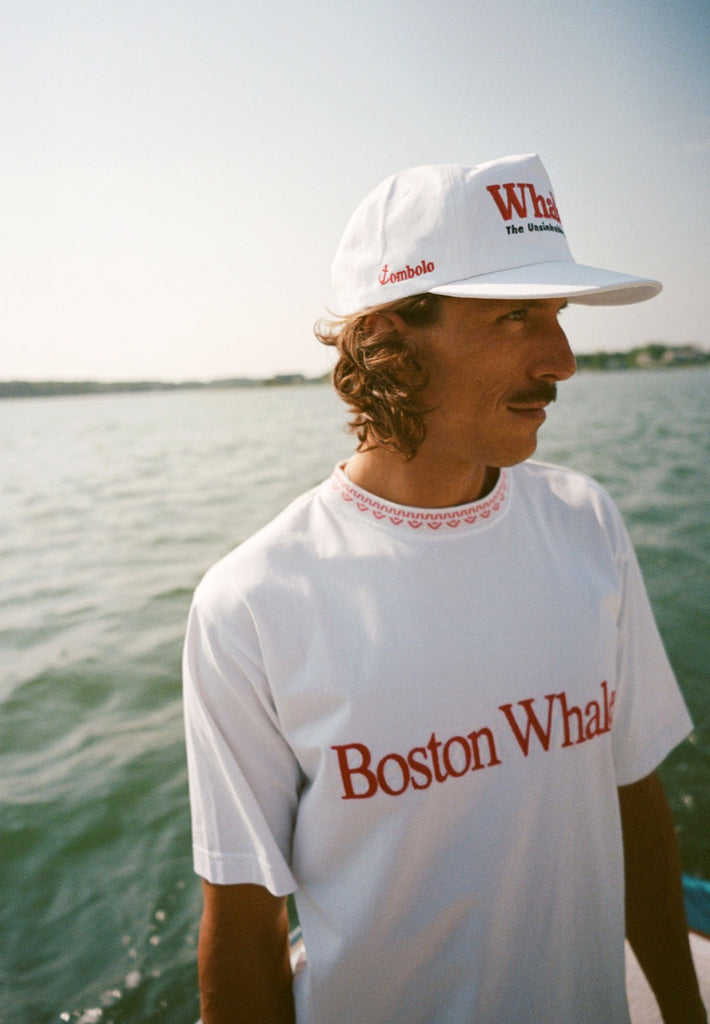 Man on a boat wearing a white 'Whaler' cap with red and black embroidery and a white T-shirt with 'Boston Whaler' in red text. The side of the cap reads 'Tombolo' in red. 