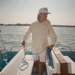 Man standing barefoot on boat deck, holding blue rope and facing sideways. He wears a cream jacket and white shorts with tan trim, along with a white cap reading 'Whaler – The Unsinkable Legend.' 