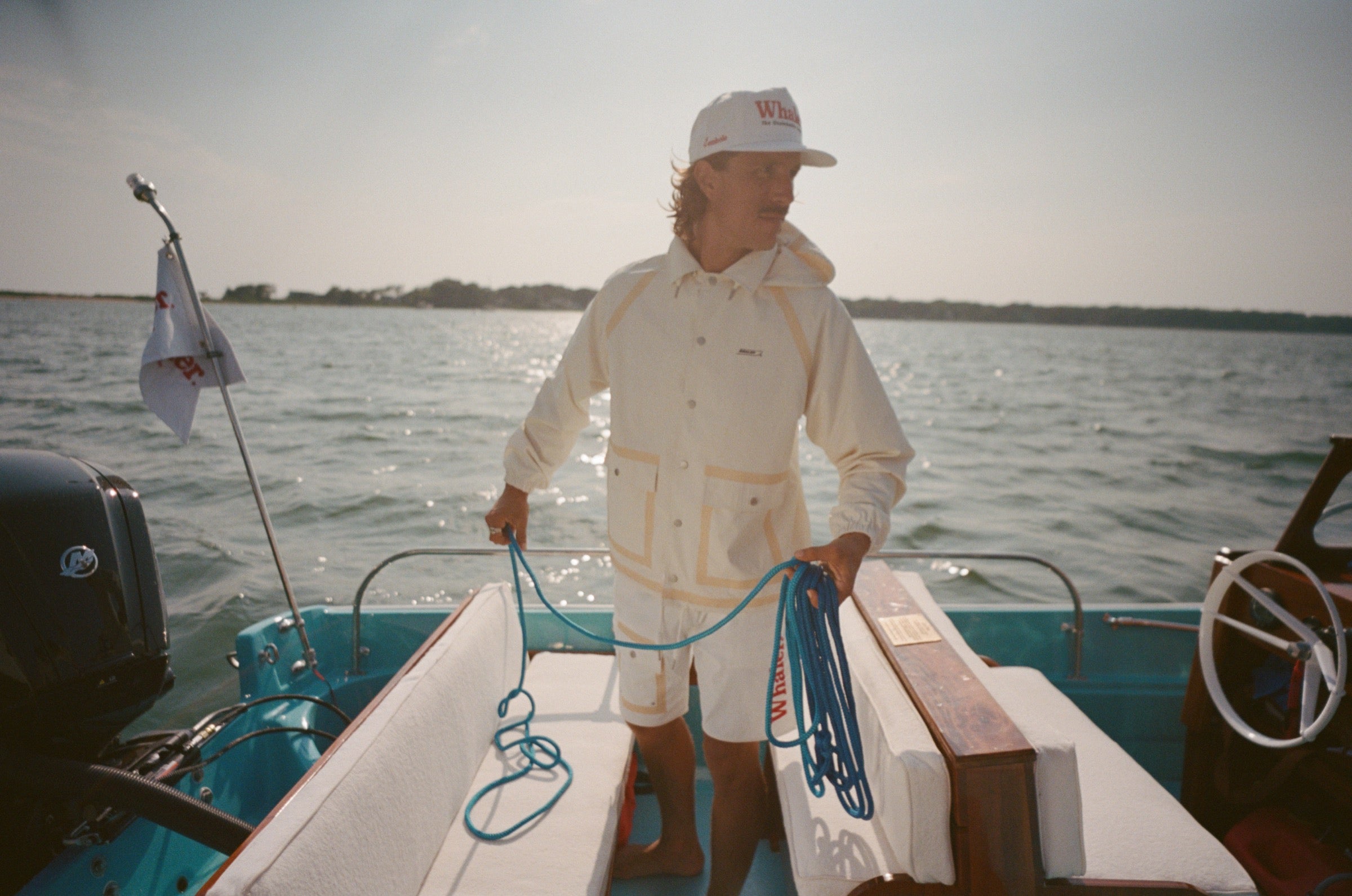 Man standing barefoot on boat deck, holding blue rope and facing sideways. He wears a cream jacket and white shorts with tan trim, along with a white cap reading 'Whaler – The Unsinkable Legend.' 