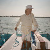 Man standing barefoot on boat deck, holding blue rope and facing sideways. He wears a cream jacket and white shorts with tan trim, along with a white cap reading 'Whaler – The Unsinkable Legend.' 