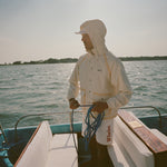 Man standing barefoot on a boat deck, holding blue rope and facing sideways. He wears a cream jacket with the hood up and matching shorts with tan trim.