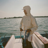Man standing barefoot on a boat deck, holding blue rope and facing sideways. He wears a cream jacket with the hood up and matching shorts with tan trim.