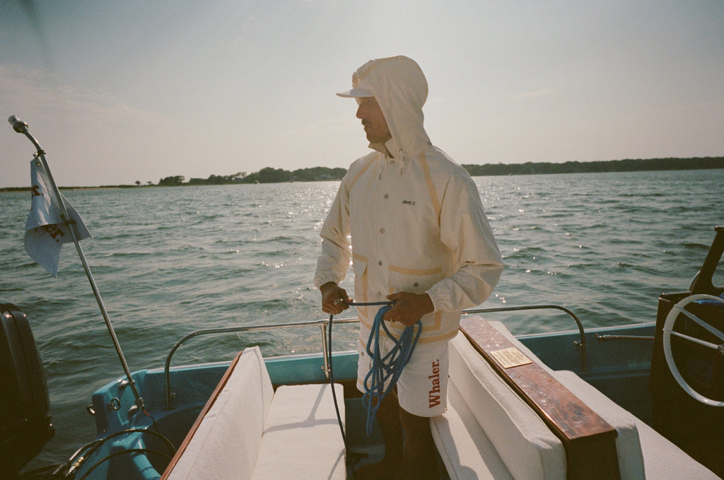 Man standing barefoot on a boat deck, holding blue rope and facing sideways. He wears a cream jacket with the hood up and matching shorts with tan trim.