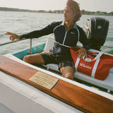 Man seated on a boat wearing a navy sun shirt with white piping and a striped collar, next to a red and white Whaler tote bag.