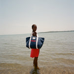 Woman standing in shallow water, holding the Navy canvas tote bag with white handles over her shoulder.