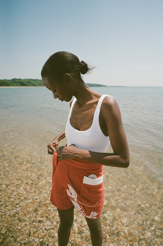 woman at beach wearing white one piece bathing suit and red skirt, looking down and tying wrap on her skirt