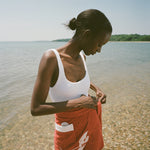 woman at beach wearing white one piece bathing suit and red skirt, looking down and tying wrap on her skirt