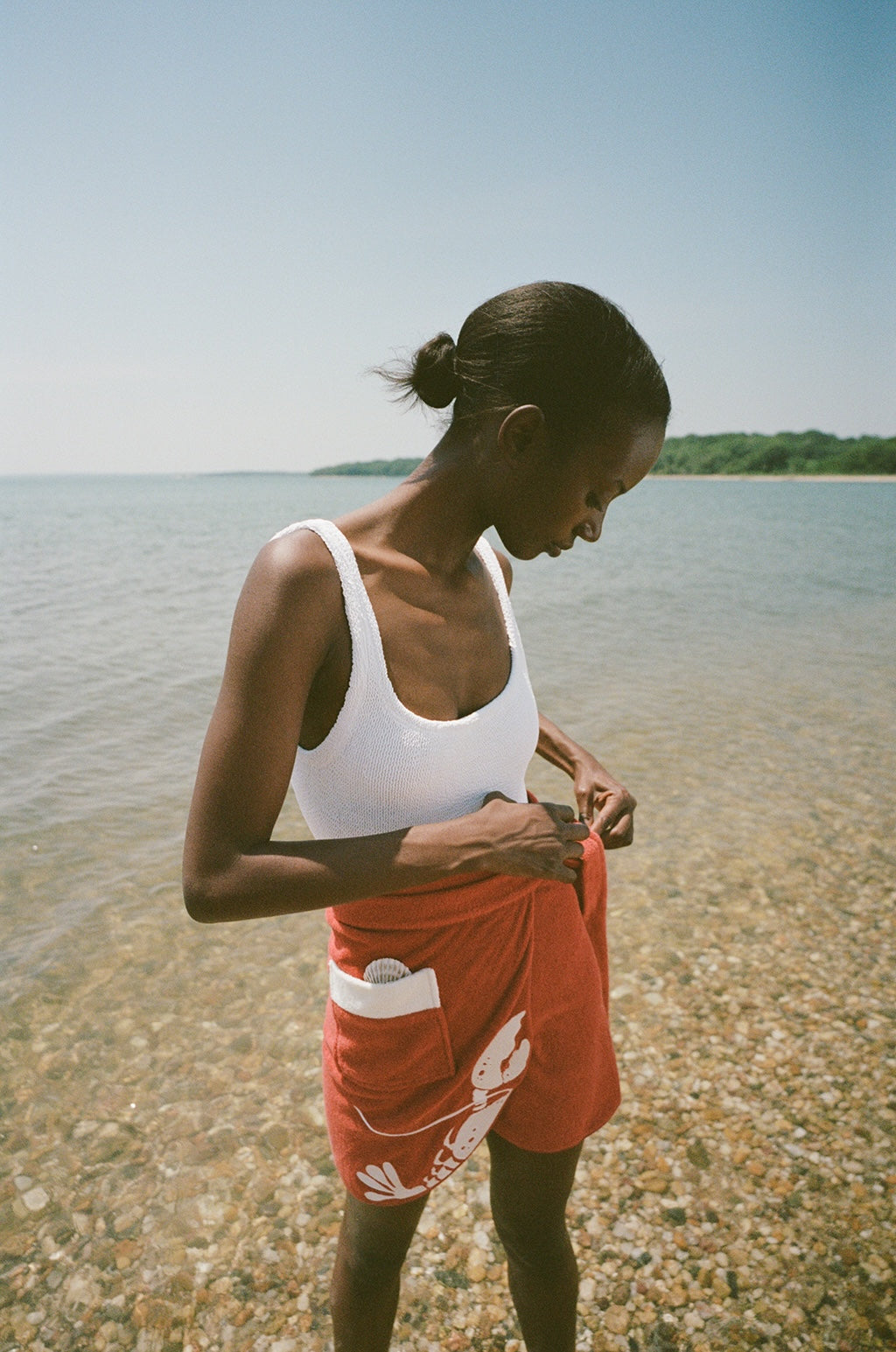 woman at beach wearing white one piece bathing suit and red skirt, looking down and tying wrap on her skirt